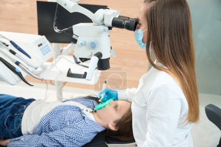 Femme dentiste traite une dent de garçon sous un microscope, elle utilise un équipement moderne