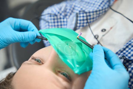 Téléchargez les photos : Jeune patient intrépide se trouve dans la chaise du dentiste, le spécialiste tient une minuscule perceuse pour nettoyer les canaux dentaires - en image libre de droit