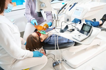 Téléchargez les photos : Adolescent dans des lunettes de protection se trouve dans la chaise du dentiste, le médecin utilise un équipement et des outils spéciaux - en image libre de droit