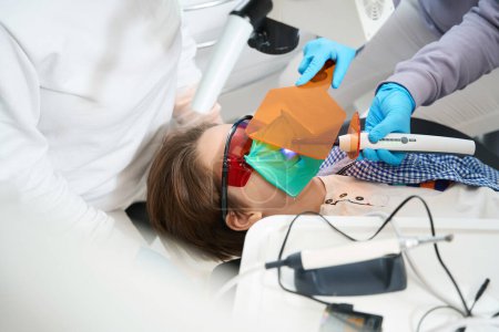 Téléchargez les photos : Garçon dans des lunettes de protection se trouve dans la chaise du dentiste, il reçoit un remplissage en polymère - en image libre de droit