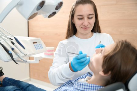 Téléchargez les photos : Garçon assis dans la chaise du dentiste, une jeune femme médecin met un sceau sur lui - en image libre de droit