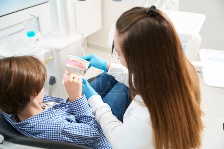 Téléchargez les photos : Garçon au cabinet de dentiste apprend les méthodes de brossage des dents et des appareils, le bureau est lumineux et propre - en image libre de droit