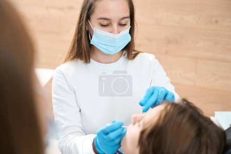 Foto de Mujer dentista en su lugar de trabajo ayuda a un niño, ella trabaja con un asistente - Imagen libre de derechos