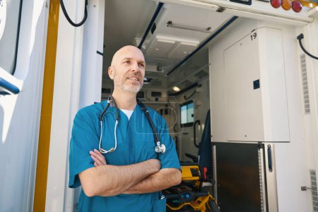 Photo for Enthusiastic doctor in a blue uniform stands with arms crossed on chest while behind him is a modern equipped saloon of medical transport - Royalty Free Image