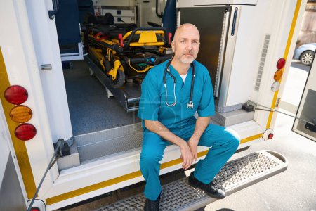 Téléchargez les photos : Un médecin fatigué en uniforme bleu est assis sur le bord d'une rampe de transport médical tandis qu'un brancard préparé pour l'hospitalisation se tient derrière lui - en image libre de droit