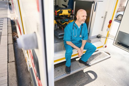 Téléchargez les photos : Un médecin joyeux en uniforme bleu est assis sur le bord d'une rampe de transport médical tandis qu'un brancard préparé pour l'hospitalisation se tient derrière lui - en image libre de droit