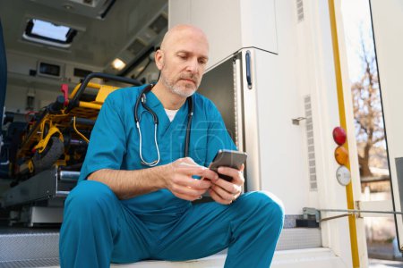 Photo for Focused doctor in blue uniform crouched on edge of medical transport ramp while looking at smartphone screen - Royalty Free Image