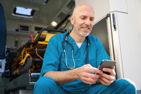 Téléchargez les photos : Médecin joyeux dans un uniforme bleu se trouve sur le bord d'une rampe de transport médical tout en regardant un écran de smartphone - en image libre de droit