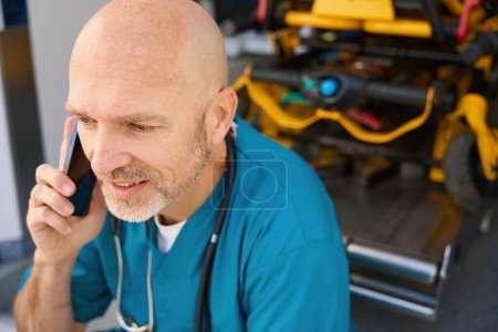Photo for Relaxed doctor in a blue uniform sits on the edge of a medical transport ramp while talking on the phone - Royalty Free Image