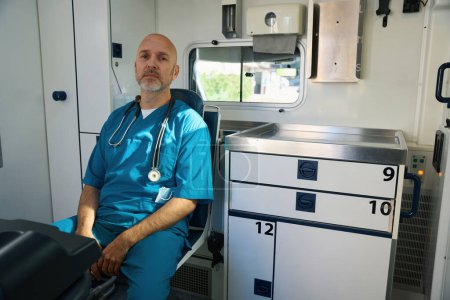 Téléchargez les photos : Médecin fatigué dans un uniforme bleu se repose alors qu'il y a des armoires avec des instruments médicaux à côté de lui - en image libre de droit