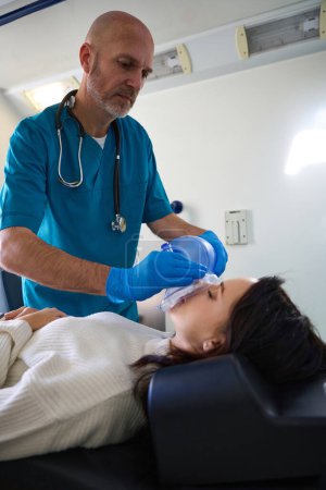 Foto de Trabajadora médica enfocada pone un dispositivo especial de ambulancia en la cara del paciente mientras ella se acuesta con los ojos cerrados inconsciente - Imagen libre de derechos