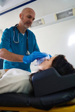 Foto de Una trabajadora médica confiada pone un dispositivo especial de ambulancia en la cara femenina mientras ella yace inconsciente con los ojos cerrados - Imagen libre de derechos