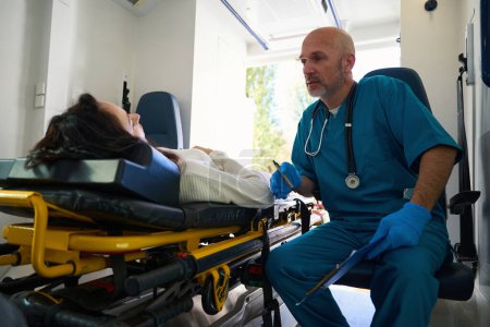 Foto de Médico atento está en un coche ambulancia mientras que una mujer hospitalizada se acuesta en una camilla - Imagen libre de derechos