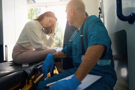Foto de Mujer triste toca su cabeza en una camilla en un vehículo médico mientras un hombre de uniforme se sienta a su lado, observando su condición - Imagen libre de derechos