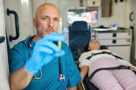 Téléchargez les photos : Un médecin concentré vérifie l "état de préparation du médicament injectable pendant qu'une femme se trouve à côté de lui sur une civière - en image libre de droit