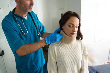 Téléchargez les photos : Un médecin soignant soigne une patiente blessée au cou alors qu'elle est assise sur un canapé - en image libre de droit
