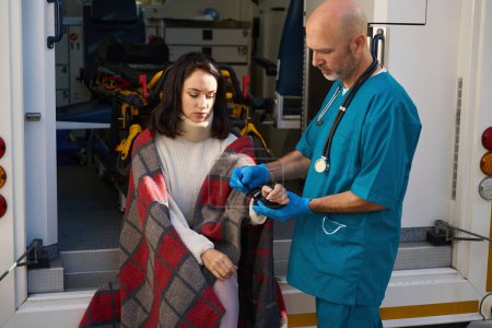 Photo for Sad patient sits wrapped in a blanket on the edge of an ambulance while a man fixes a fixing bandage on his wrist - Royalty Free Image