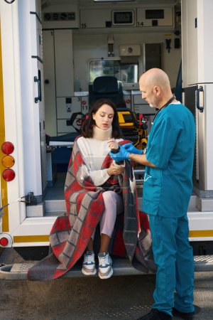 Foto de La frustrada paciente se sienta envuelta en una manta con el brazo levantado mientras el hombre le pone un aparato médico en la muñeca. - Imagen libre de derechos
