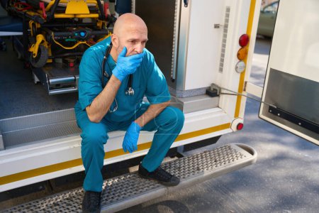 Photo for Sad man in a medical uniform and sterile gloves sits near a mobile medical equipment while pain is displayed on his face - Royalty Free Image