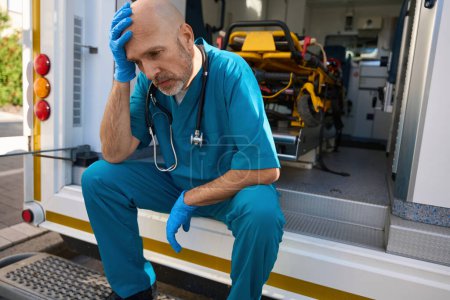 Téléchargez les photos : Un homme surmené dans un uniforme médical est assis sur le bord d'une voiture médicale tandis que sa main touche sa tête - en image libre de droit
