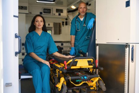 Téléchargez les photos : Heureux ambulancier d'une ambulance touche l'équipement dans un transport pour l'hospitalisation des patients tandis que son collègue dans des gants stériles se tient à proximité - en image libre de droit