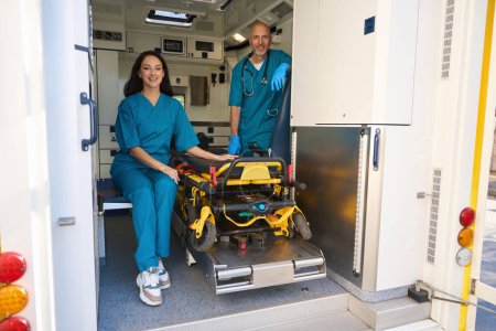 Photo for Happy paramedic from an ambulance crew sits near a stretcher in a transport for hospitalization of patients while her colleague in sterile gloves stands nearby - Royalty Free Image