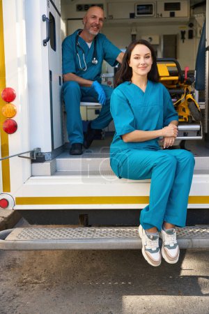 Téléchargez les photos : Femme satisfaite dans un uniforme médical se trouve près d'une civière dans le transport d'ambulance tandis qu'un homme accroupi à proximité dans des gants stériles - en image libre de droit