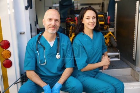 Photo for Happy ambulance woman sits on the edge of a medical transport while her colleague sits next to her in sterile gloves on her hands - Royalty Free Image