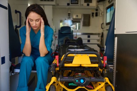 Foto de Enfermera agotada agachada en el asiento junto a la camilla en el interior del coche para atención médica urgente - Imagen libre de derechos