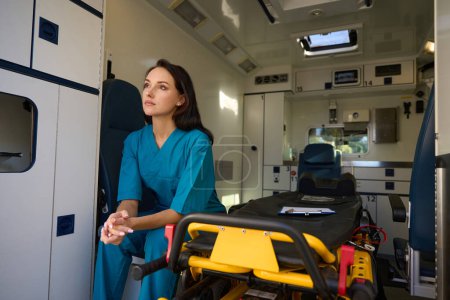 Foto de Enfermera pensativa sentada en un asiento junto a una camilla en el interior de un automóvil para atención médica de emergencia - Imagen libre de derechos