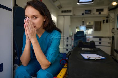 Téléchargez les photos : Triste infirmière s'assoit sur un siège à côté d'une civière dans une voiture d'urgence tandis que ses mains sont pliées devant son visage - en image libre de droit