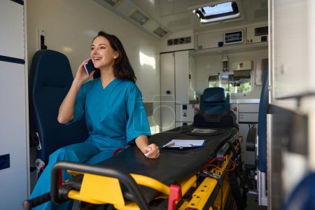 Téléchargez les photos : Infirmière joyeuse assise sur un siège à côté d'une civière à l'intérieur d'une voiture pour une assistance médicale d'urgence lors d'une conversation téléphonique - en image libre de droit
