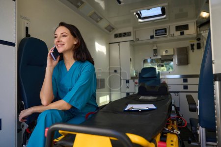 Foto de Enfermera alegre sentada en un asiento junto a una camilla en el interior de un coche para atención médica urgente durante una conversación telefónica - Imagen libre de derechos