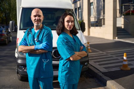 Photo for Confident man and woman in medical uniform standing next to friend with crossed arms over chest while resuscitation vehicle parked behind them - Royalty Free Image