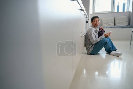 Téléchargez les photos : Portrait de vue latérale de joyeuse femme gaie est assis sur le sol tout en tenant le téléphone portable dans la main dans l'appartement moderne - en image libre de droit