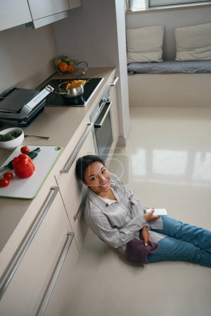 Foto de Mujer africana está sentada en el suelo mientras mira y posa en la cámara fotográfica en la moderna cocina blanca - Imagen libre de derechos