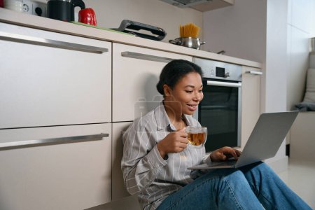 Foto de Cintura hacia arriba retrato de la mujer africana positiva feliz con la taza de té en la mano está escribiendo algo en el ordenador portátil en el apartamento de casa moderna - Imagen libre de derechos