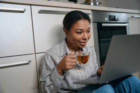 Foto de Primer plano retrato de hermosa hembra sonriente con taza de té en la mano durante el trabajo en línea desde el apartamento en casa - Imagen libre de derechos