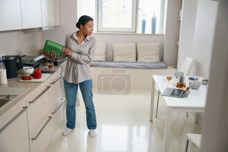 Téléchargez les photos : Portrait complet de la femme élégante dans la cuisine tout en faisant le travail à la maison tout en regardant l'écran de l'ordinateur portable - en image libre de droit