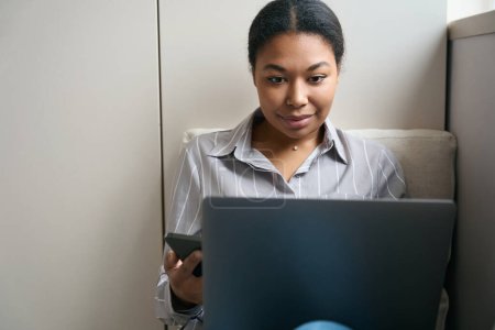 Foto de Retrato de vista frontal de la encantadora dama de negocios africana está utilizando el ordenador portátil mientras trabaja en línea desde casa - Imagen libre de derechos