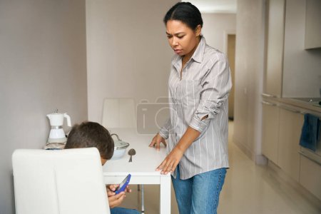 Téléchargez les photos : Vue de côté portrait de la mère bouleversée debout près du garçon dans la cuisine alors qu'il joue sur l'appareil moderne - en image libre de droit