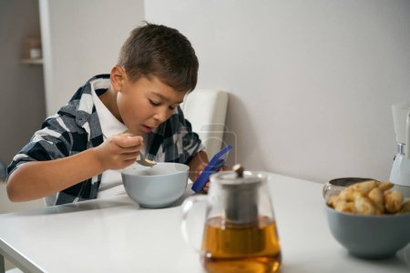 Photo for Waist up portrait of handsome Caucasian male child eating breakfast while holding game console in hand in the kitchen - Royalty Free Image