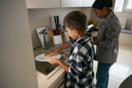 Téléchargez les photos : Tour de taille arrière portrait d'un garçon d'âge scolaire aide sa mère dans la cuisine moderne - en image libre de droit