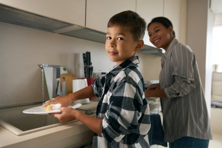 Foto de Primer plano retrato de guapo caucásico un escuela envejecido chico es ayudar a madre con un housework en la cocina - Imagen libre de derechos