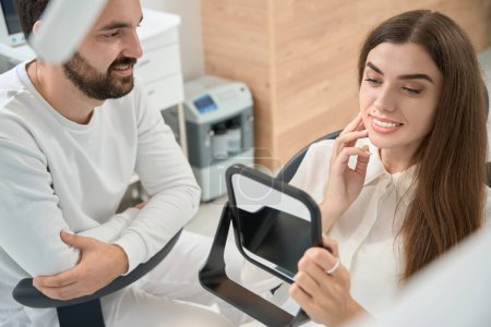 Téléchargez les photos : Heureuse cliente de clinique dentaire regardant dans un miroir de main en présence de son stomatologue - en image libre de droit