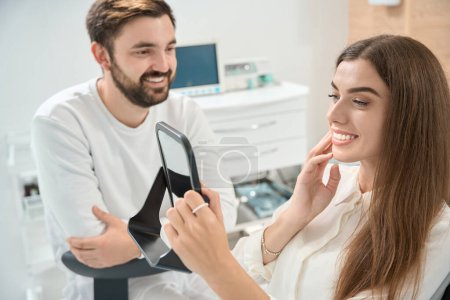Photo for Joyful female patient looking at herself in hand mirror in presence of happy stomatologist - Royalty Free Image