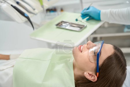 Téléchargez les photos : Patiente calme dans un bavoir jetable et des lunettes de sécurité allongées sur une chaise pendant que son dentiste choisit un instrument de diagnostic - en image libre de droit