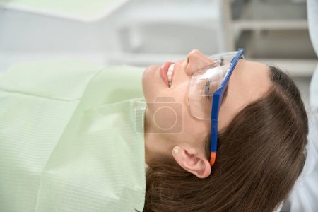 Photo for Smiling female patient in disposable bib and safety goggles lying in chair in dentist office - Royalty Free Image