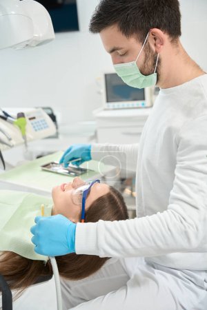 Photo for Dentist looking at calm patient lying in dental chair while picking out dental instrument - Royalty Free Image