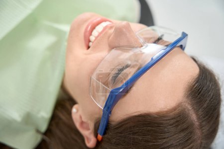 Photo for Pleased client in protective eyewear and disposable bib lying supine in chair in dentist office before examination - Royalty Free Image